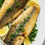 Two cooked fish fillets on a white platter with lemon slices and herbs.