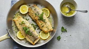 Two cooked swordfish fillets in a skillet with lemon slices, herbs, and a side of sauce.