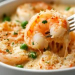 Close-up of Garlic shrimp gratin in a white bowl, shrimp on a fork with melted cheese, breadcrumbs, parsley, and peas visible. Creamy texture.