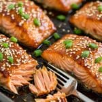 Flaky baked teriyaki salmon fillets on a dark pan, garnished with sesame seeds and green onions. Fork tender.