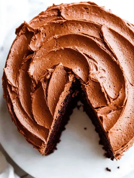Close-up of a chocolate cake with creamy frosting and a slice cut out revealing soft, moist layers.