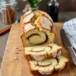 Sliced Amish cinnamon bread on a wooden board, showcasing the swirl. Cinnamon sticks and a bottle in the background.