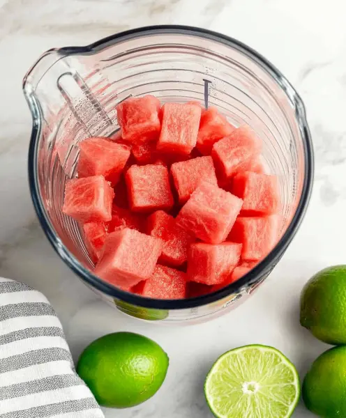 Cubed watermelon in a measuring cup with fresh whole and halved limes nearby.