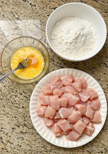 Diced raw chicken pieces, a bowl of whisked eggs, and a bowl of all-purpose flour, ready for breading.
