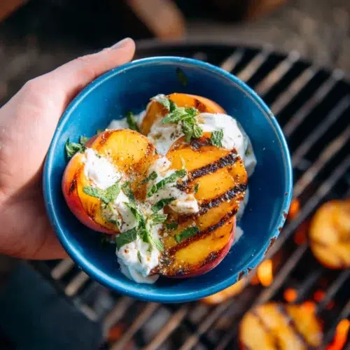 Grilled peach halves with char marks served in a blue bowl with honey yogurt and mint, held above a flaming grill.