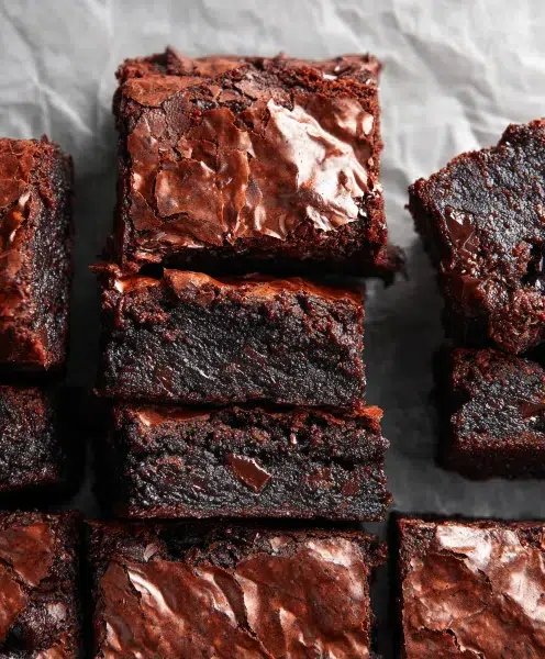 Fudgy brownie squares stacked on parchment paper with shiny crackly tops and rich chocolate interiors