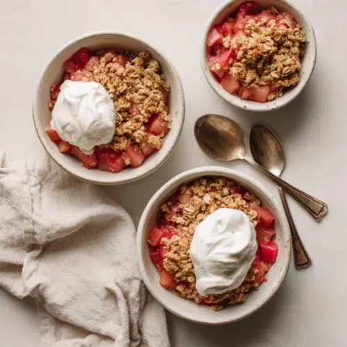 Bowls of rhubarb crisp topped with golden crumble and whipped cream, served on a light countertop with spoons.