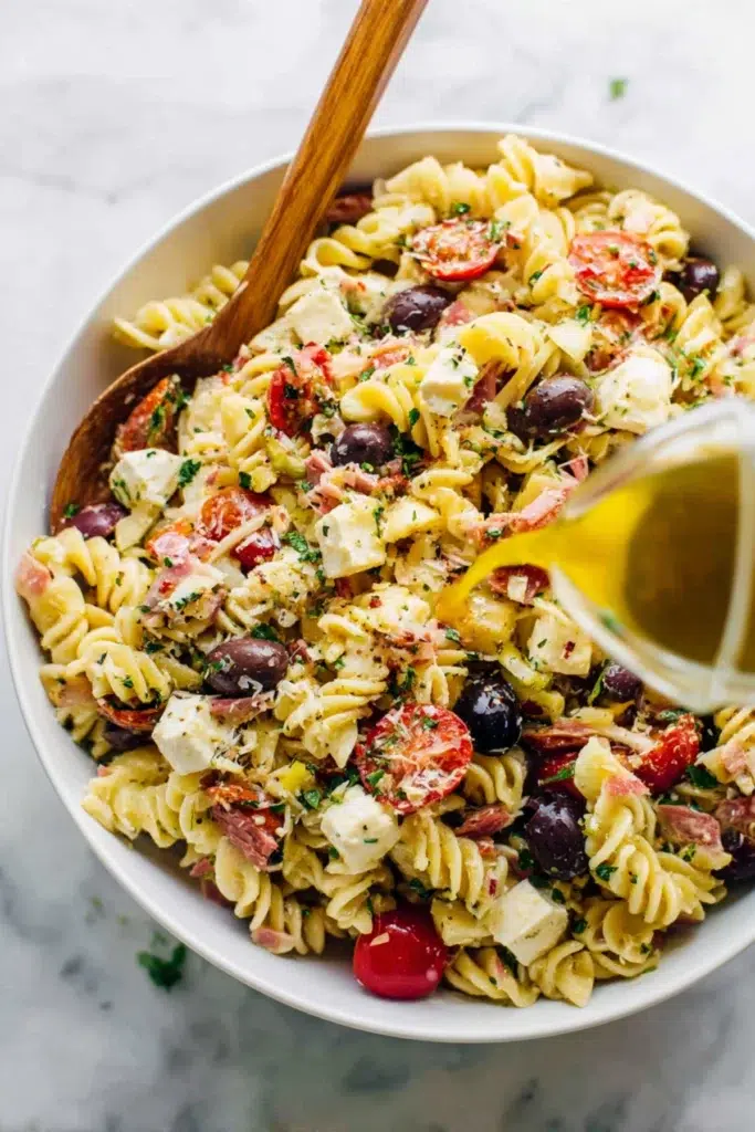 Italian pasta salad with rotini, cherry tomatoes, olives, feta, and herbs being dressed with olive oil in a white bowl.