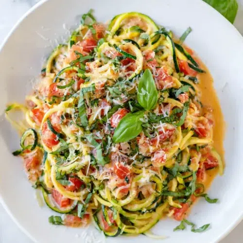 Fresh zucchini zoodle pasta with cherry tomatoes, parmesan cheese, and basil served in a white bowl.