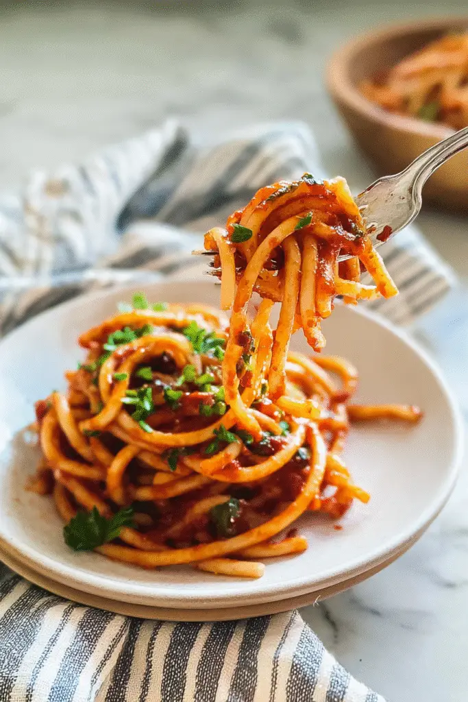 Twirl of caramelized shallot pasta on a fork with rich tomato sauce and fresh herbs on a white plate