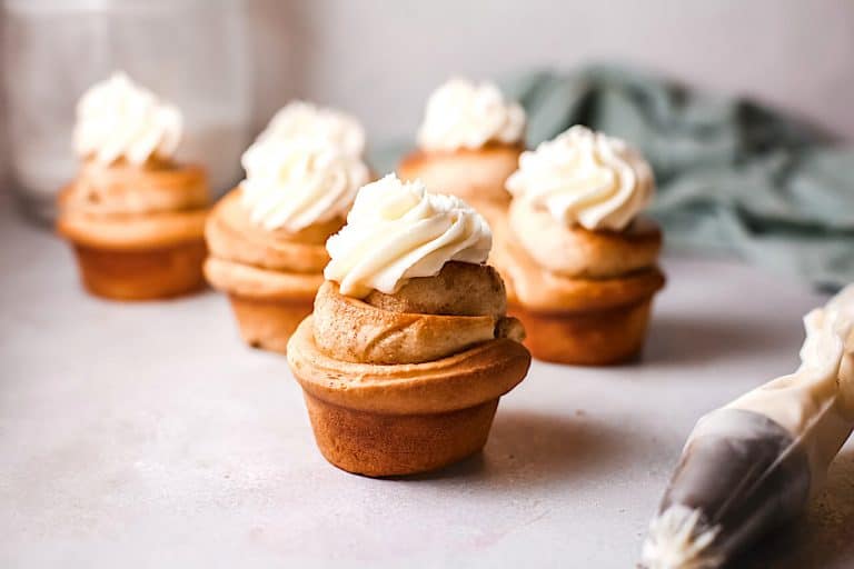 Golden brown cinnamon roll cupcakes topped with creamy frosting swirls, piping bag nearby on a light background