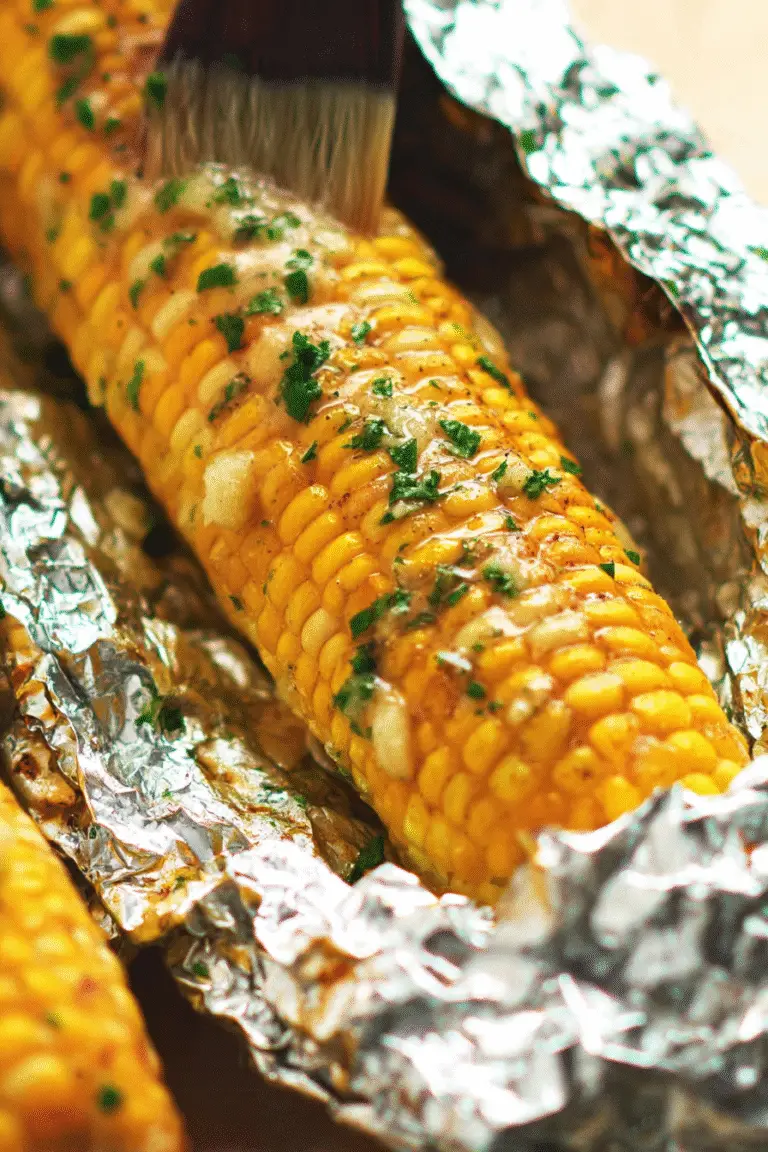 Close-up of oven-roasted corn on the cob in foil, brushed with garlic herb butter and sprinkled with fresh parsley