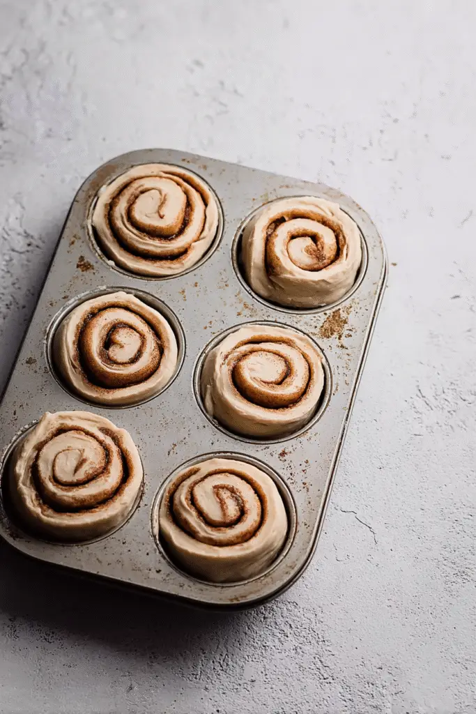 Unbaked cinnamon roll dough spirals in a muffin tin, ready for baking into golden, fluffy cinnamon roll cupcakes