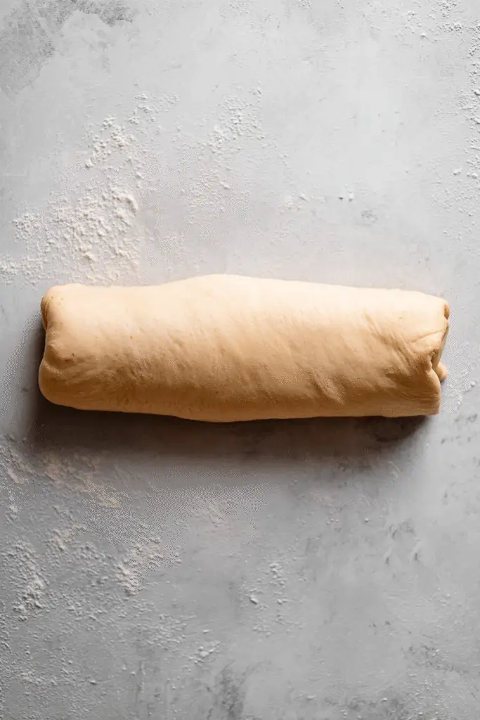 Top-down view of rolled cinnamon roll dough log on a floured surface, ready to be sliced into individual cupcake portions