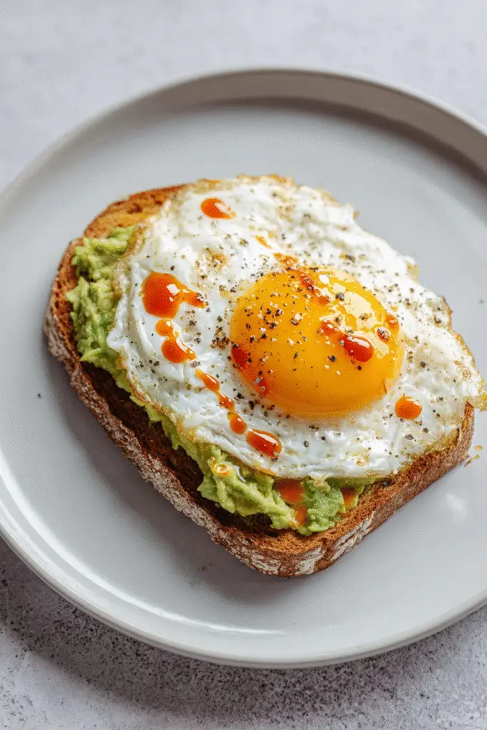 Golden fried egg with runny yolk on avocado toast, seasoned with pepper and drizzled with hot sauce for extra flavor