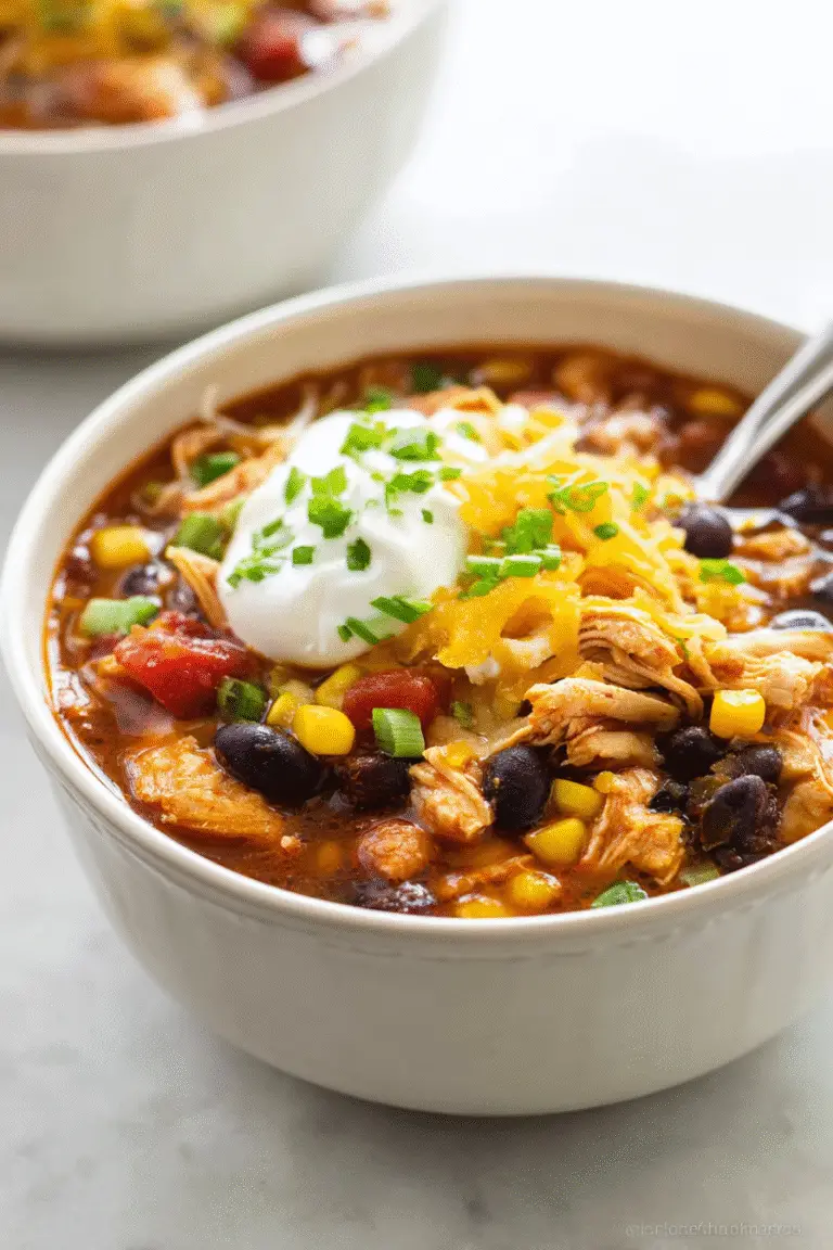 Hearty chicken taco soup with beans, corn, tomatoes, sour cream, cheddar cheese, and green onions in a white bowl.