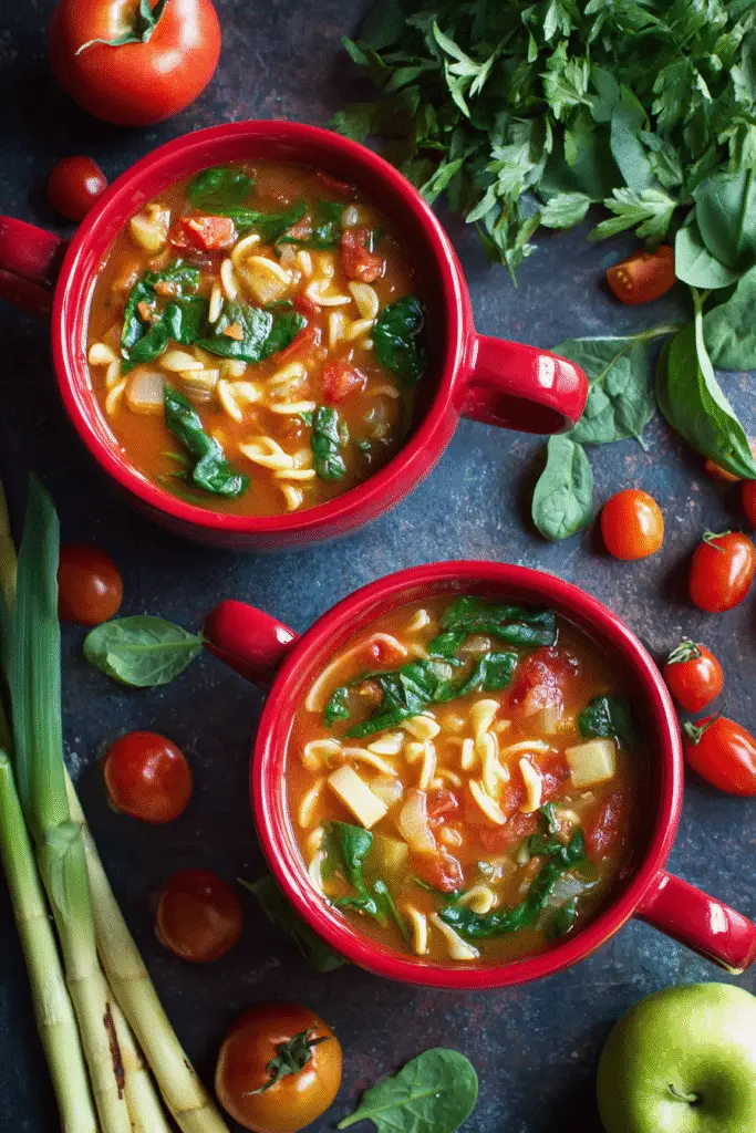 Warm bowl of vegetable orzo soup with spinach, tomatoes, and pasta served in red mugs on a rustic kitchen table